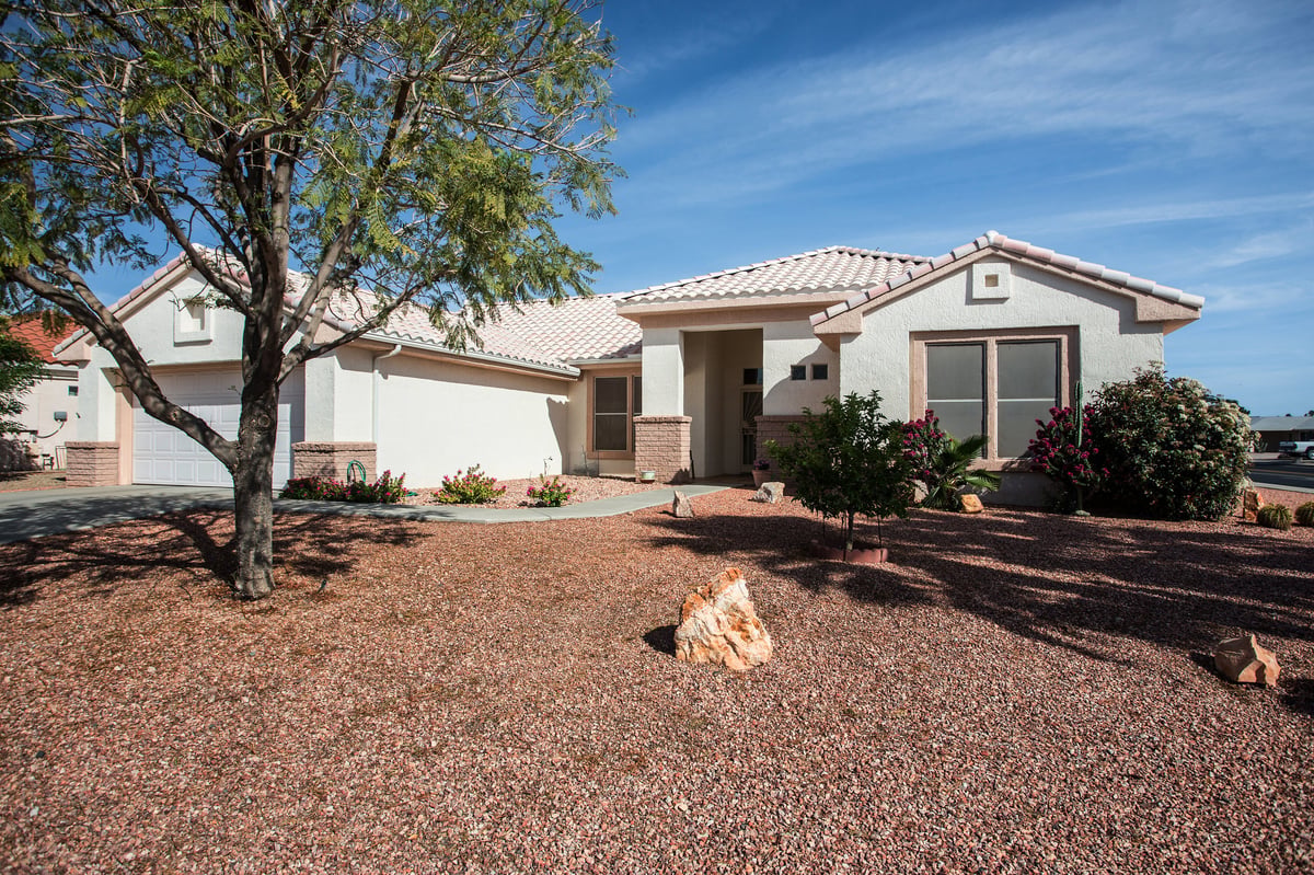 Single-story beige stucco house with tile roof in desert landscape, featuring rock yard, large tree, and clear blue sky