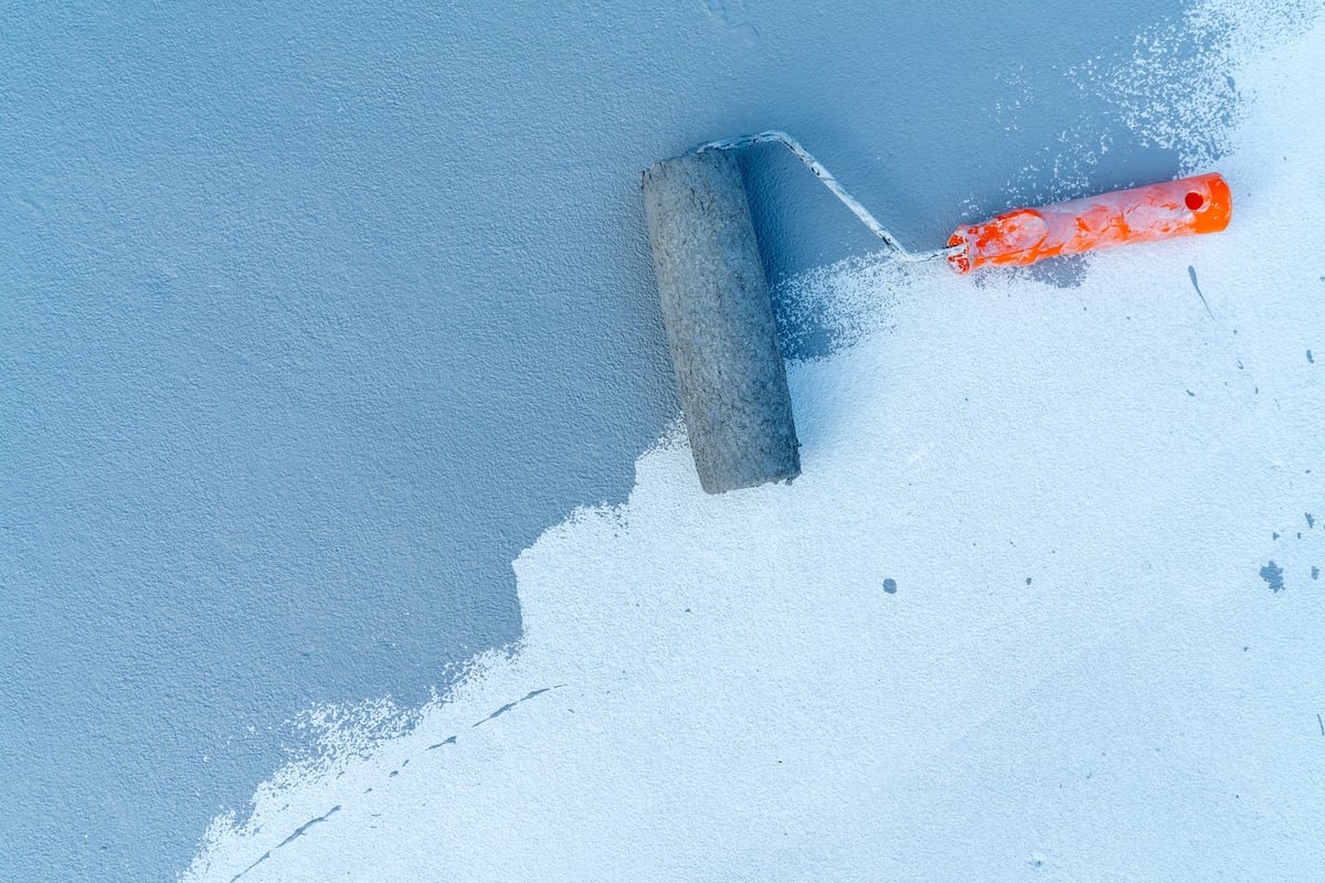 Paint roller with orange handle applying blue paint on a textured white concrete surface